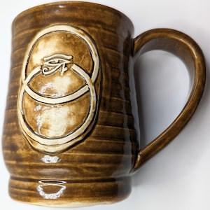 Op de afbeelding: Een bruine keramische mok met een wit ontwerp van een oog in twee overlappende cirkels. De mok heeft een handvat en een getextureerd, gestreept oppervlak.