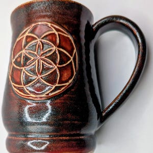 May include: A handmade brown ceramic mug with a large handle. The mug features a raised, circular design of interlocking circles and petals. The mug has a glossy finish and is suitable for hot or cold beverages.
