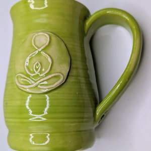 Puede incluir: Una taza de cerámica verde lima hecha a mano con un asa grande. La taza presenta un diseño en relieve blanquecino de una persona en postura de yoga en la parte delantera. La taza tiene un acabado brillante y una superficie texturizada y acanalada.