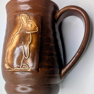 May include: A brown ceramic mug with a large handle. The mug features a raised relief of a rabbit within a rectangular frame. The mug has a textured surface and is suitable for hot beverages.