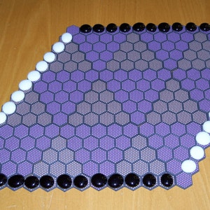 Peut inclure: Un plateau de jeu hexagonal violet avec des pièces de jeu noires et blanches autour du périmètre.