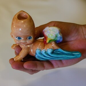 Könnte beinhalten: Eine Keramikfigur eines Babys, das auf einer Welle reitet, mit einer Regenbogenwolke dahinter. Die Figur hat ein Loch oben auf dem Kopf, was darauf hindeutet, dass es sich um eine Pfeife handelt.