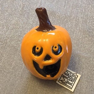 May include: A ceramic orange pumpkin with a smiling face and a brown stem. The pumpkin has black eyes and a black mouth. A small tag with the text "MUK HETZ" is attached.