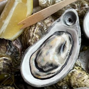 May include: An overhead shot of fresh oysters in their shells, arranged on a metal tray. A lemon wedge and a wooden fork are placed near the oysters. The oyster shells are a mix of gray, white, and brown.