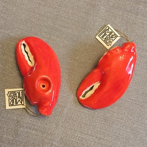 May include: Two glossy red ceramic lobster claw ornaments with white interior details. Each piece features a small circular opening and a white square tag attached with a gold string, printed with the text Mud Hedz in a triangular logo design.