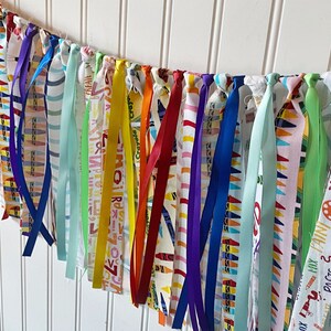May include: A colorful fabric banner with a variety of ribbons and fabric scraps. The fabric is printed with a crayon pattern and the words "Crayola Crayon", "MIX PATNI DESIGN FIIX", "PASTE", "&Sigma;", "Аяла", "RIBBLE PASTE", "BRUSH SCRIBBLE", "JICHT", and "CREATE".