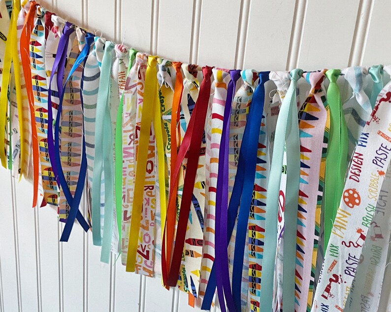 May include: A colorful fabric banner with a variety of ribbons and fabric scraps. The fabric is printed with a crayon pattern and the words "Crayola Crayon", "MIX PATNI DESIGN FIIX", "PASTE", "&Sigma;", "Аяла", "RIBBLE PASTE", "BRUSH SCRIBBLE", "JICHT", and "CREATE".