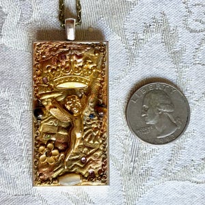 K&ouml;nnte beinhalten: Rechteckiger, goldfarbener Anh&auml;nger mit einem erhabenen Reliefdesign mit einem Cherub, einer Krone und floralen Elementen. Der Anh&auml;nger ist an einer Kette befestigt. Eine M&uuml;nze liegt neben dem Anh&auml;nger zum Gr&ouml;&szlig;envergleich.