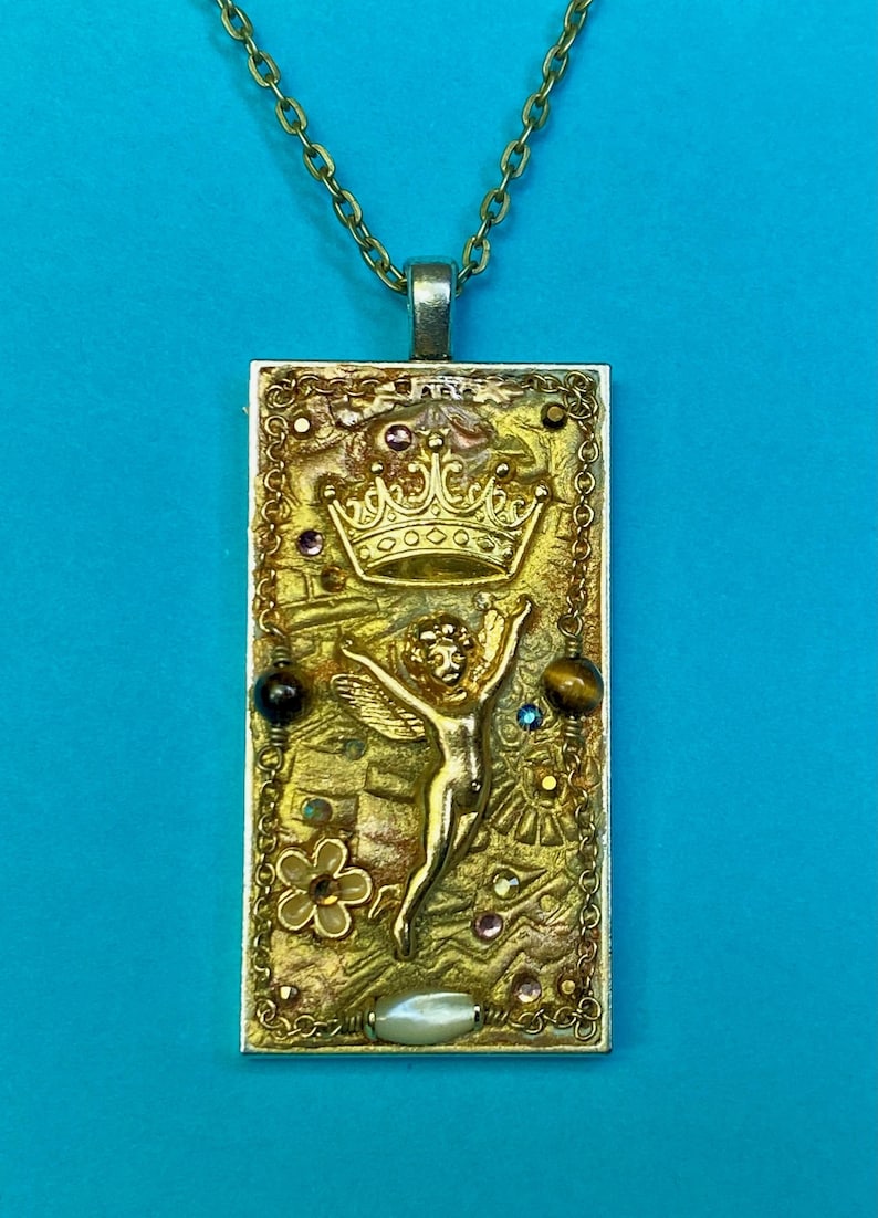 K&ouml;nnte beinhalten: Goldfarbene Anh&auml;nger Halskette mit einem rechteckigen Anh&auml;nger mit Krone, Engel und verschiedenen Edelsteinen. Der Anh&auml;nger hat eine strukturierte Oberfl&auml;che und h&auml;ngt an einer filigranen Kette. Der Hintergrund ist blau.