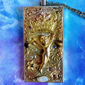 K&ouml;nnte beinhalten: Rechteckiger Anh&auml;nger mit goldfarbenem Rahmen und detailliertem Design. Der Anh&auml;nger zeigt eine goldene Krone, eine kleine Figur mit ausgestreckten Armen und verschiedene Verzierungen, darunter Edelsteine und eine Blume. Er h&auml;ngt an einer Kette.