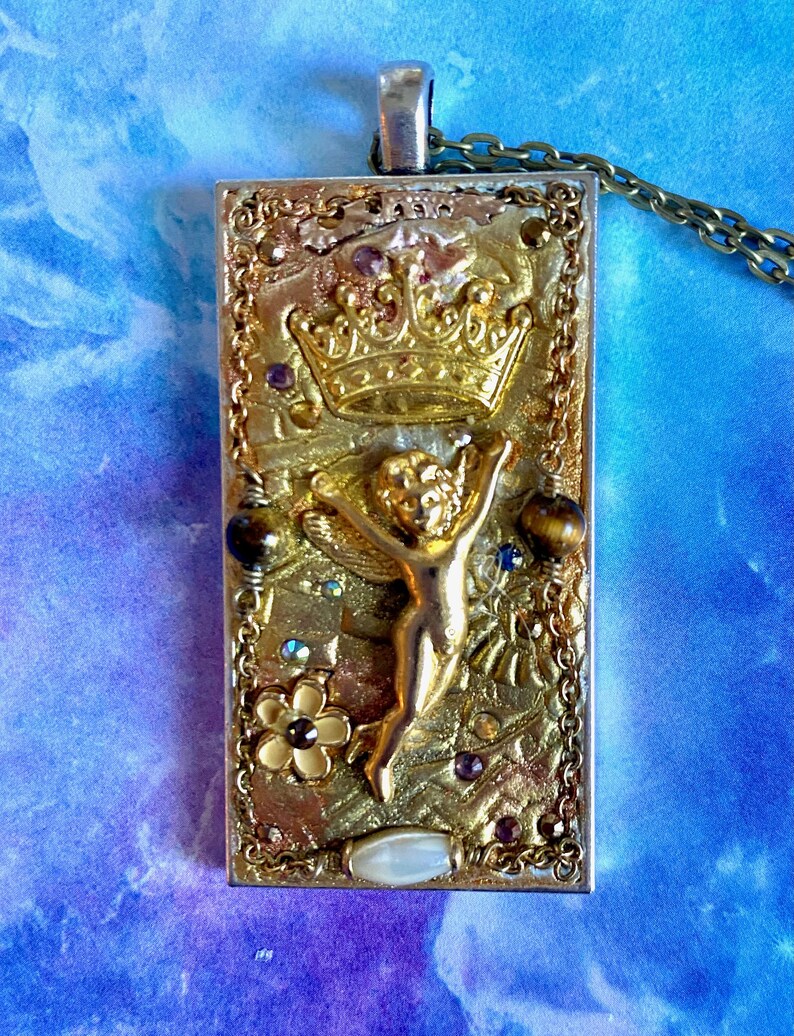 K&ouml;nnte beinhalten: Rechteckiger Anh&auml;nger mit goldfarbenem Rahmen und detailliertem Design. Der Anh&auml;nger zeigt eine goldene Krone, eine kleine Figur mit ausgestreckten Armen und verschiedene Verzierungen, darunter Edelsteine und eine Blume. Er h&auml;ngt an einer Kette.