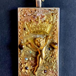 K&ouml;nnte beinhalten: Goldfarbene rechteckige Anh&auml;nger-Halskette mit detailliertem Design. Der Anh&auml;nger zeigt eine Krone, einen Cherub und florale Elemente, akzentuiert mit braunen und irisierenden Edelsteinen. Die Halskette hat eine zarte Kette.