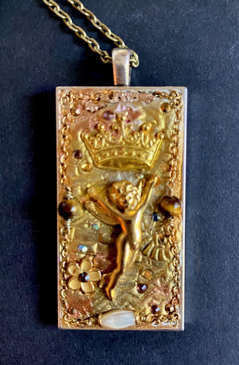 K&ouml;nnte beinhalten: Goldfarbene rechteckige Anh&auml;nger-Halskette mit detailliertem Design. Der Anh&auml;nger zeigt eine Krone, einen Cherub und florale Elemente, akzentuiert mit braunen und irisierenden Edelsteinen. Die Halskette hat eine zarte Kette.