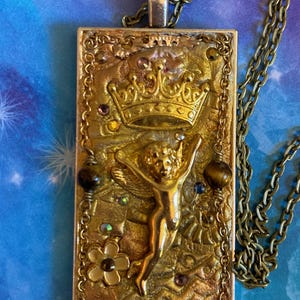 K&ouml;nnte beinhalten: Rechteckige Anh&auml;nger-Halskette mit goldfarbener Kette. Der Anh&auml;nger zeigt ein goldfarbenes Reliefdesign einer Krone, eines Engels und floraler Elemente, akzentuiert mit farbigen Edelsteinen. Der Anh&auml;nger ist ca. 5 cm lang.
