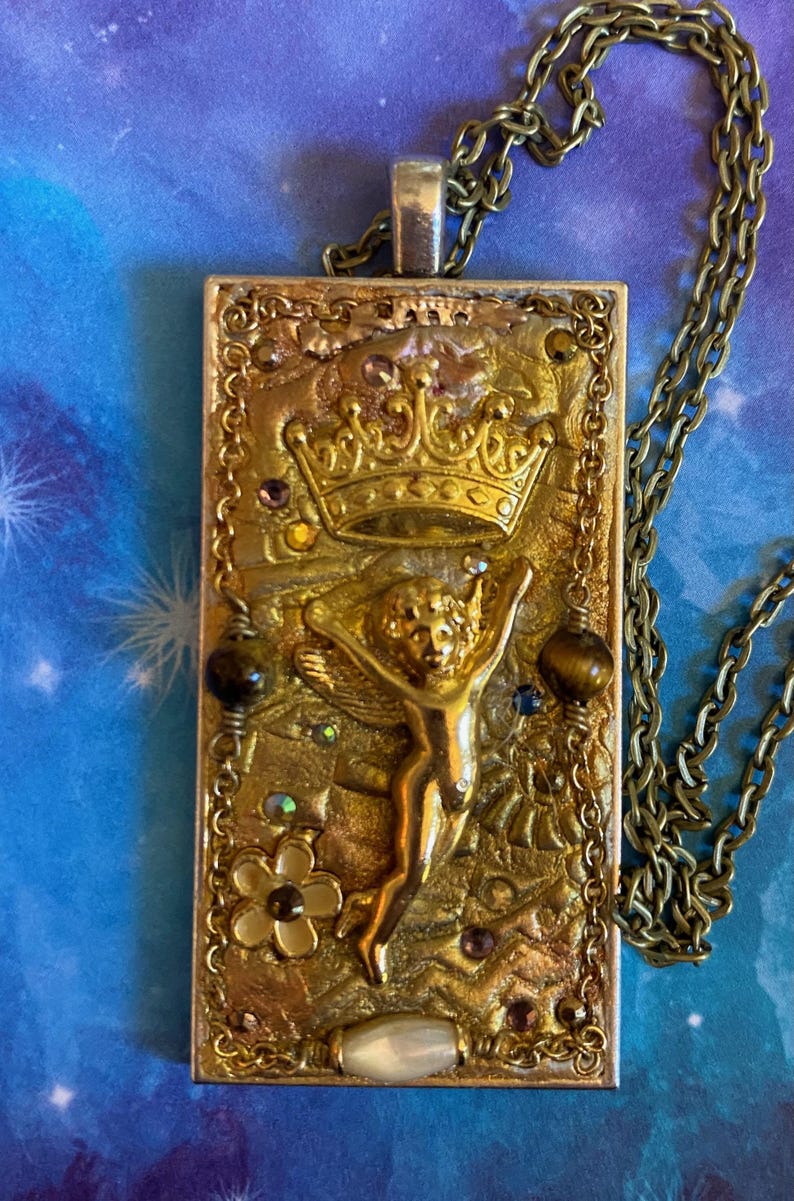 K&ouml;nnte beinhalten: Rechteckige Anh&auml;nger-Halskette mit goldfarbener Kette. Der Anh&auml;nger zeigt ein goldfarbenes Reliefdesign einer Krone, eines Engels und floraler Elemente, akzentuiert mit farbigen Edelsteinen. Der Anh&auml;nger ist ca. 5 cm lang.