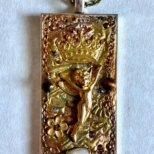 K&ouml;nnte beinhalten: Rechteckiger Anh&auml;nger mit goldfarbenem Rahmen und detailliertem Design. Der Anh&auml;nger zeigt eine goldene Krone, eine kleine Figur mit erhobenen Armen und verschiedene Verzierungen, darunter kleine Edelsteine und eine Blume. Er h&auml;ngt an einer filigranen Kette.