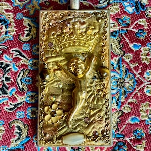 K&ouml;nnte beinhalten: Eine goldfarbene Anh&auml;ngerkette mit einem rechteckigen Anh&auml;nger. Der Anh&auml;nger zeigt ein erhabenes Relief eines Cherubs mit einer Krone, umgeben von dekorativen Elementen und kleinen Edelsteinen. Der Hintergrund ist ein gemustertes rot-blaues Gewebe.