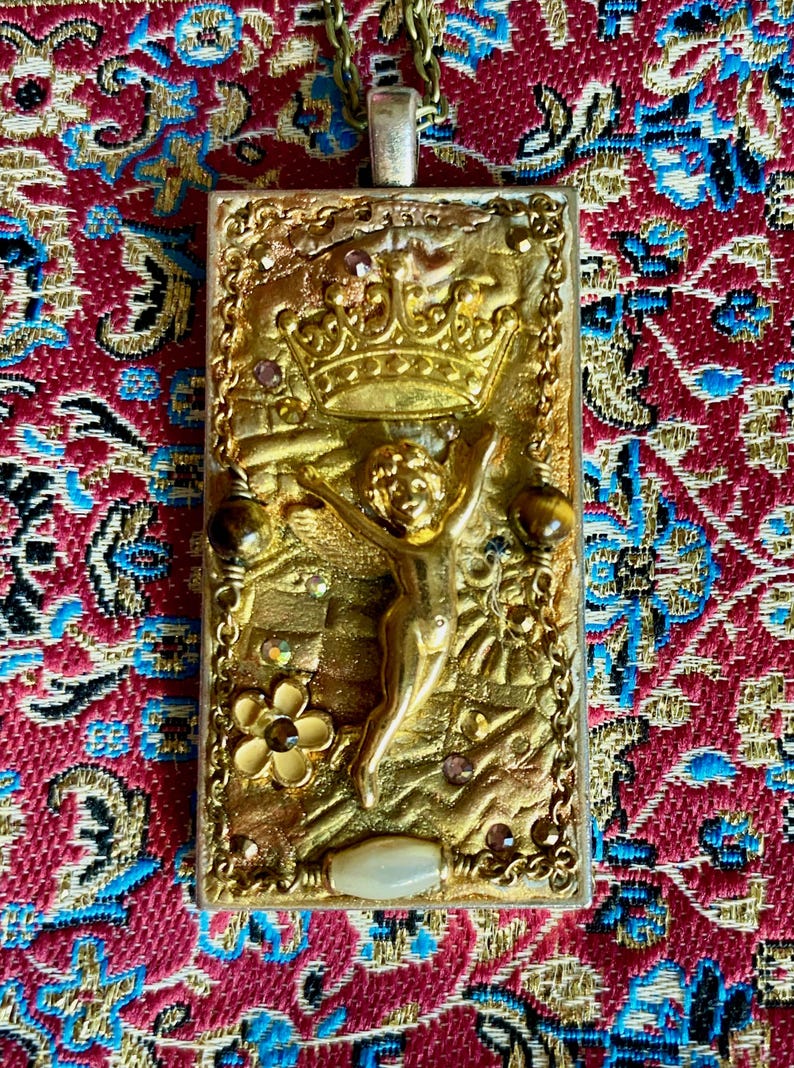 K&ouml;nnte beinhalten: Eine goldfarbene Anh&auml;ngerkette mit einem rechteckigen Anh&auml;nger. Der Anh&auml;nger zeigt ein erhabenes Relief eines Cherubs mit einer Krone, umgeben von dekorativen Elementen und kleinen Edelsteinen. Der Hintergrund ist ein gemustertes rot-blaues Gewebe.