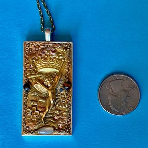 K&ouml;nnte beinhalten: Eine goldfarbene rechteckige Anh&auml;ngerhalskette mit detailliertem Design. Der Anh&auml;nger zeigt eine Krone, eine Engelsfigur und verschiedene Verzierungen. Die Halskette befindet sich an einer Kette, und eine M&uuml;nze befindet sich neben dem Anh&auml;nger zum Gr&ouml;&szlig;envergleich.