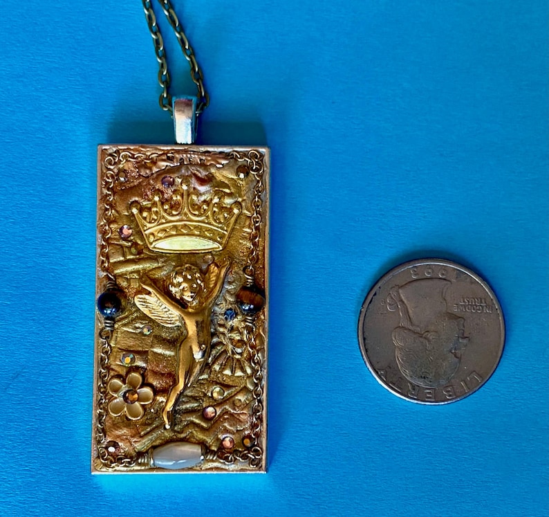 K&ouml;nnte beinhalten: Eine goldfarbene rechteckige Anh&auml;ngerhalskette mit detailliertem Design. Der Anh&auml;nger zeigt eine Krone, eine Engelsfigur und verschiedene Verzierungen. Die Halskette befindet sich an einer Kette, und eine M&uuml;nze befindet sich neben dem Anh&auml;nger zum Gr&ouml;&szlig;envergleich.