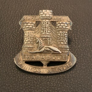 Könnte beinhalten: Ein silbernes Metallabzeichen mit einem Schlossdesign und dem Text "SEMPER FIDELIS" über dem Schloss. Der Text "MARABOUT PRIMUS IN INDIS" befindet sich unter dem Schloss.