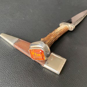 May include: A small knife with a textured brown antler handle and a silver bolster featuring a large, square-cut orange gemstone. The knife is accompanied by a brown leather sheath with silver metal caps at both ends, resting in a horizontal position.