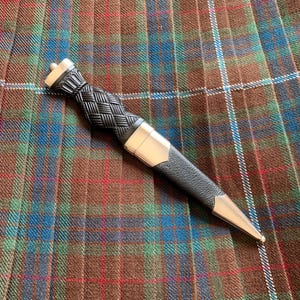 Peut inclure: Un sgian-dubh avec un manche noir texturé au motif tressé et un pommeau argenté. Le fourreau est noir avec des garnitures métalliques argentées sur le haut et une pointe effilée, posé sur un tissu à carreaux marron, vert et bleu.