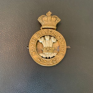 May include: A vintage brass badge with the words "NORTH STAFFORDSHIRE" encircling a central emblem. The emblem features a crown above a design with three plumes. The badge has a pin on the back.
