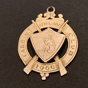 May include: A gold-coloured metal pendant with a shield design featuring a person holding a rifle. The shield is surrounded by two crossed rifles and a banner that reads "CARNEGIE RIFLE CLUB 1906".