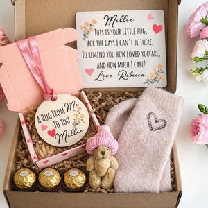 May include: A gift box containing a pink box, a wooden ornament, a pair of socks, a teddy bear, and three chocolate candies. The ornament says "A HUG FROM ME TO YOU Millie". A card reads "Millie, This is your little hug...Love Rebecca".