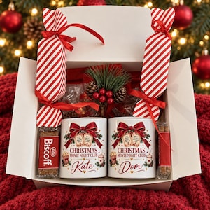 May include: A white gift box containing two ceramic mugs with the text "Christmas Movie Night Club" and the names "Kate" and "Dom". The box includes two red and white striped candy-shaped packages, a Biscoff biscuit, and a pinecone decoration.