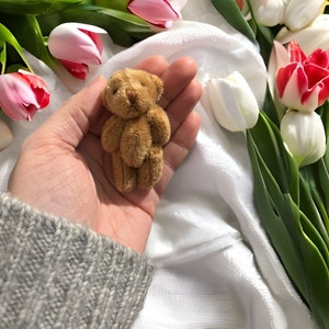 May include: A small, brown teddy bear with a soft, plush texture. The bear is held in a hand, with a background of white fabric and colorful tulips. The bear is a classic toy.