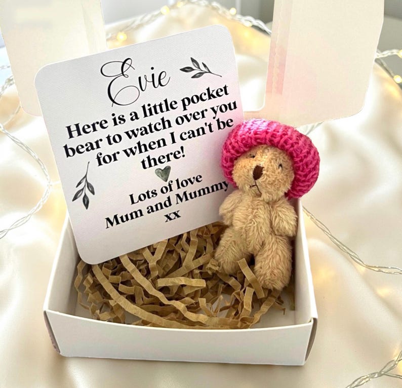 May include: A small, plush teddy bear wearing a pink knitted hat, presented in a white gift box with shredded paper. A card reads "Evie, Here is a little pocket bear to watch over you for when I can't be there! Lots of love Mum and Mummy XX."