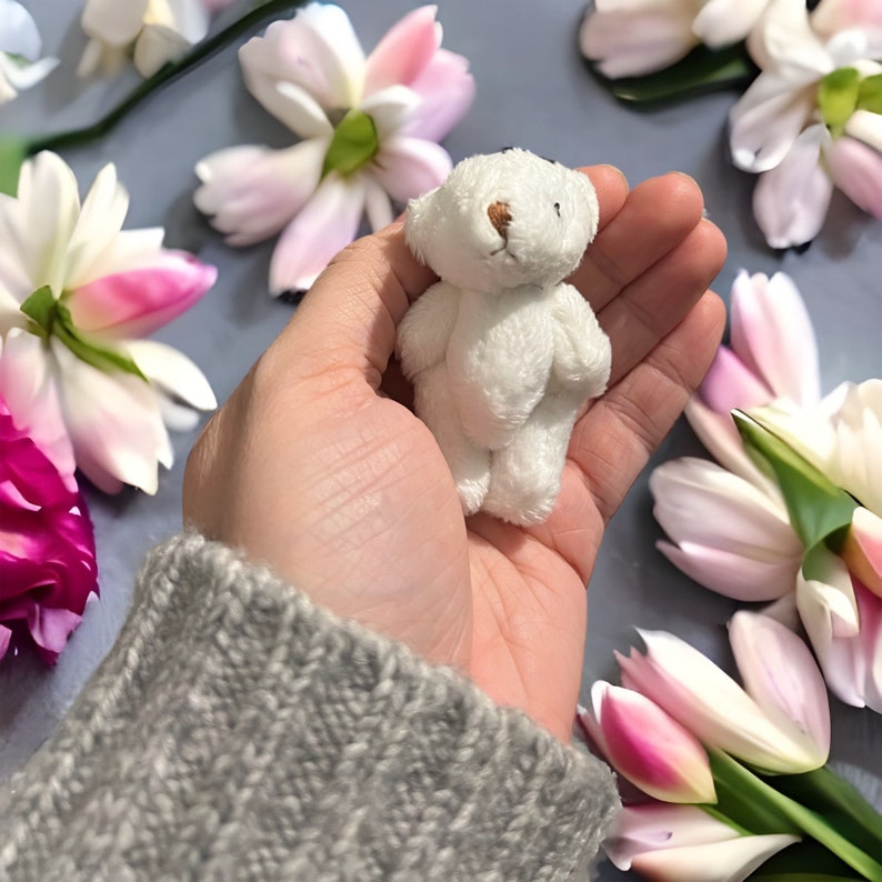 May include: A small, white teddy bear with a light brown nose and small black eyes. The plush toy is held in a hand, surrounded by pink and white flowers. The bear is approximately 2 inches tall.