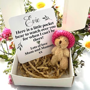 May include: A small, tan teddy bear wearing a pink knitted hat, nestled in a white box filled with shredded paper. A card with the name "Evie" and a message from "Mum and Mummy" is propped up in the box. The box is surrounded by flowers.