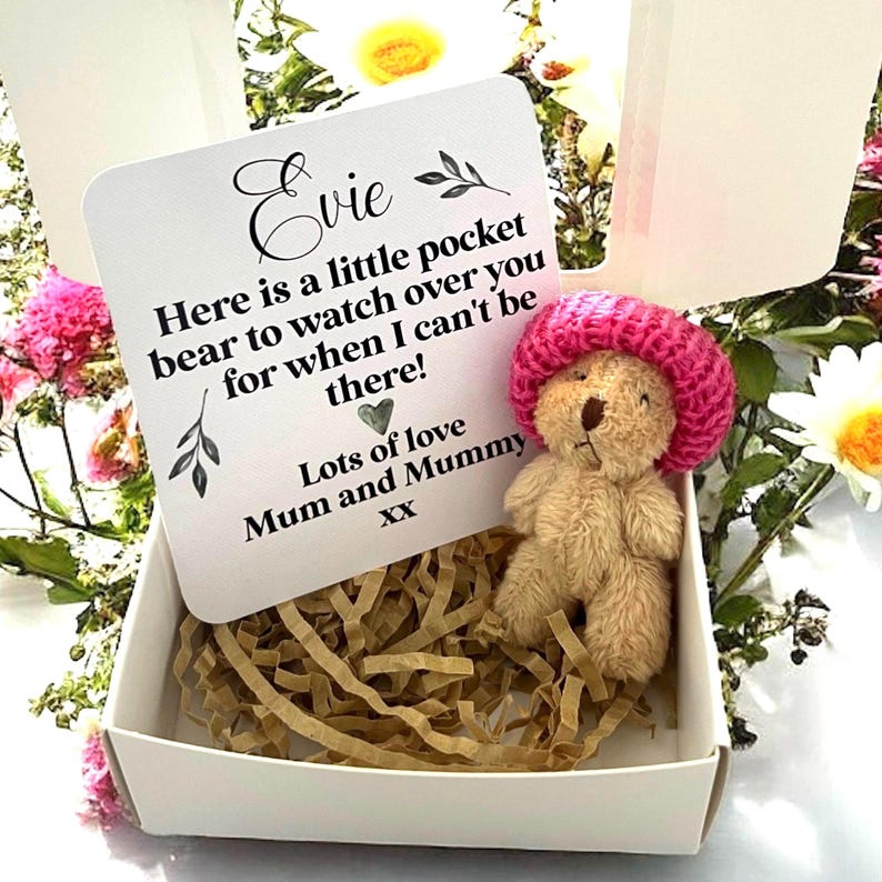 May include: A small, tan teddy bear wearing a pink knitted hat, nestled in a white box filled with shredded paper. A card with the name "Evie" and a message from "Mum and Mummy" is propped up in the box. The box is surrounded by flowers.