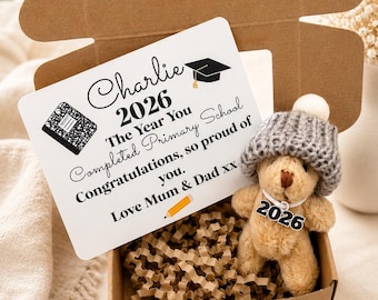 Op de afbeelding: Een afstudeergeschenkdoos met een teddybeer met een grijze gebreide muts en een label met "2026". De doos bevat een kaart met "Charlie 2026 The Year You Completed Primary School". De doos is gevuld met bruin papier.