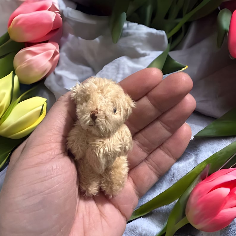 May include: A small, tan teddy bear with a stitched nose and black eyes. The bear is held in a hand, surrounded by pink and yellow tulips. The bear is a miniature, likely a collectible or decorative item.