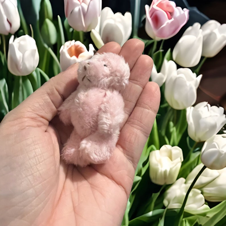 May include: A small, pink teddy bear made of soft, plush material. The bear is held in a hand, with a backdrop of white and pink tulips. The bear has a simple design with visible stitching and a small, rounded head.