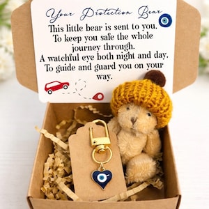 May include: A small teddy bear wearing a yellow knitted hat is in a brown box with shredded paper. A gold keychain with a blue heart-shaped charm and an evil eye design hangs beside the bear. The box contains a card with the text "Your Protection Bear".