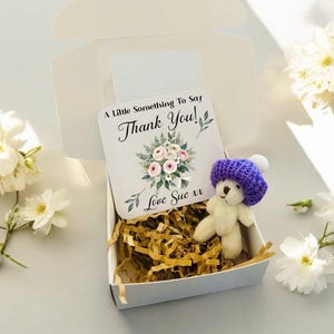 May include: A white gift box contains a small teddy bear wearing a purple knitted hat, a card with floral design and the text "Thank You!", and decorative paper filler. White flowers are arranged around the box.