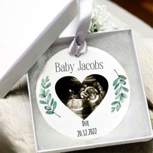 May include: White ceramic bauble with a heart-shaped ultrasound image, green leaves, and the text "Baby Jacobs" and "DUE 20.12.2022".