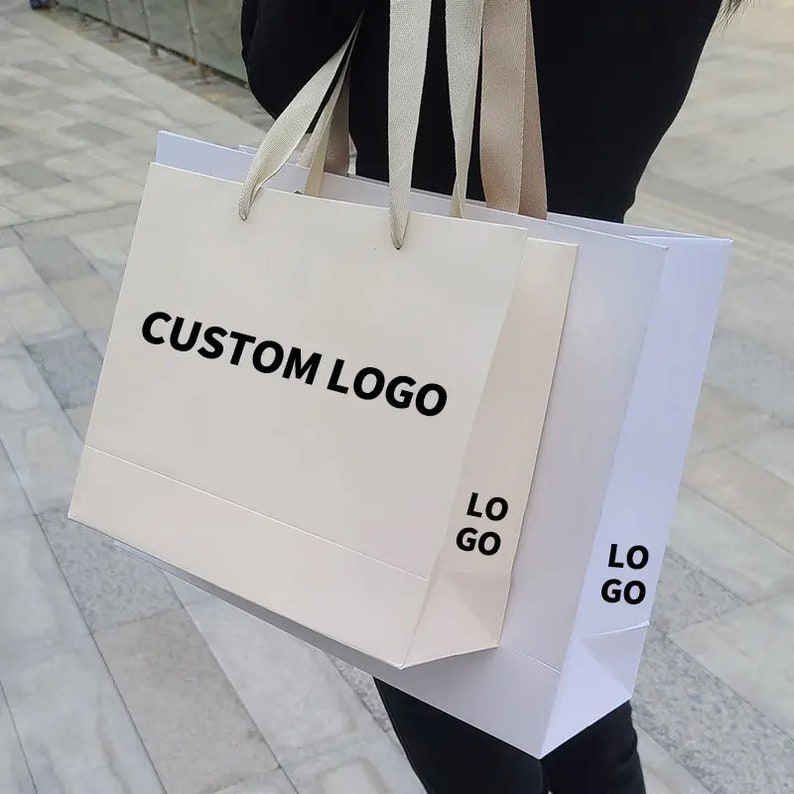 May include: Two white shopping bags with cream-colored ribbon handles. The larger bag has the text "CUSTOM LOGO" printed in black. The smaller bag has the text "LOGO" printed in black.