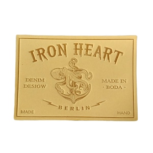 May include: A rectangular, tan leather patch with embossed text and a nautical design. The words "IRON HEART" are arched across the top. Below, it reads "DENIM DESIGN MADE IN BODA HAND BERLIN". An octopus is over an anchor.