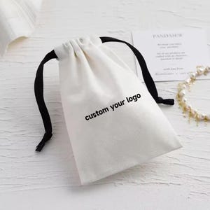 Könnte beinhalten: Ein weißer Kordelzugbeutel mit schwarzen Kordeln. Auf der Tasche steht "custom your logo". Eine Karte und eine Perlenkette befinden sich im Hintergrund.