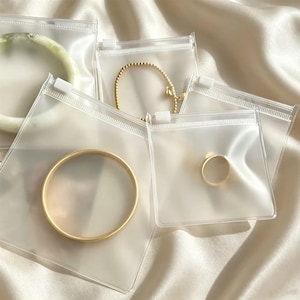 May include: A collection of jewelry pieces individually packaged in clear, square plastic zipper bags. The items include a gold-toned beaded bracelet, a smooth gold-toned bangle, a simple gold-toned ring, and a pale green circular bangle.