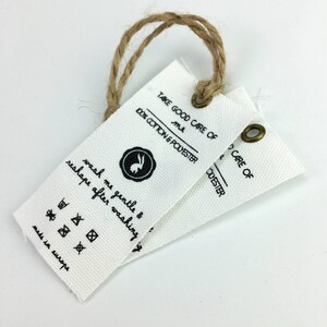 May include: Two white fabric tags with black text and a small bunny logo. The tags are attached with a brown twine string. The text on the tags reads "Take good care of me 100% cotton & polyester" and "wash me gentle & reshape after washing".