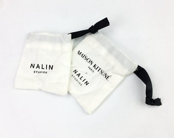 May include: Two white cotton drawstring bags with black drawstrings. The bag on the left has the text "NALIN STUDIOS" printed on it. The bag on the right has the text "MAISON KITSUNÉ PARIS" and "NALIN STUDIOS" printed on it.