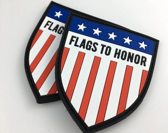Puede incluir: Dos parches bordados en rojo, blanco y azul con forma de escudo. Los parches tienen un borde negro y el texto "FLAGS TO HONOR" en letras negras.