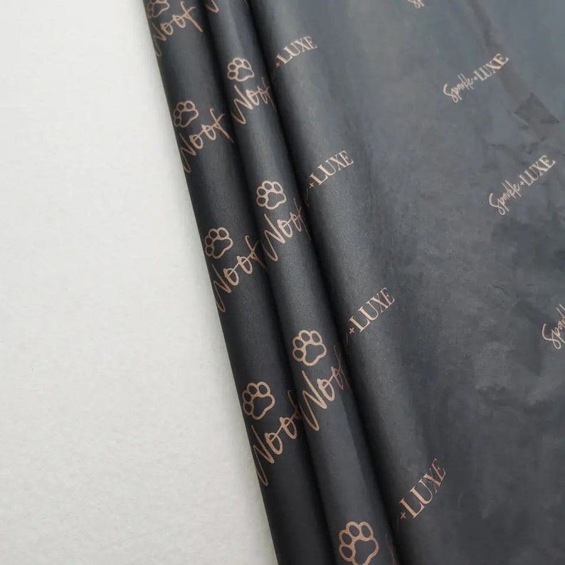 K&ouml;nnte beinhalten: Schwarzes Seidenpapier mit einem sich wiederholenden Muster aus Pfotenabdr&uuml;cken und den Worten "Woof Noof" und "Sparkle Luxe" in metallischem Ros&eacute;gold. Das Papier ist gerollt und zeigt seine Textur und sein Design.
